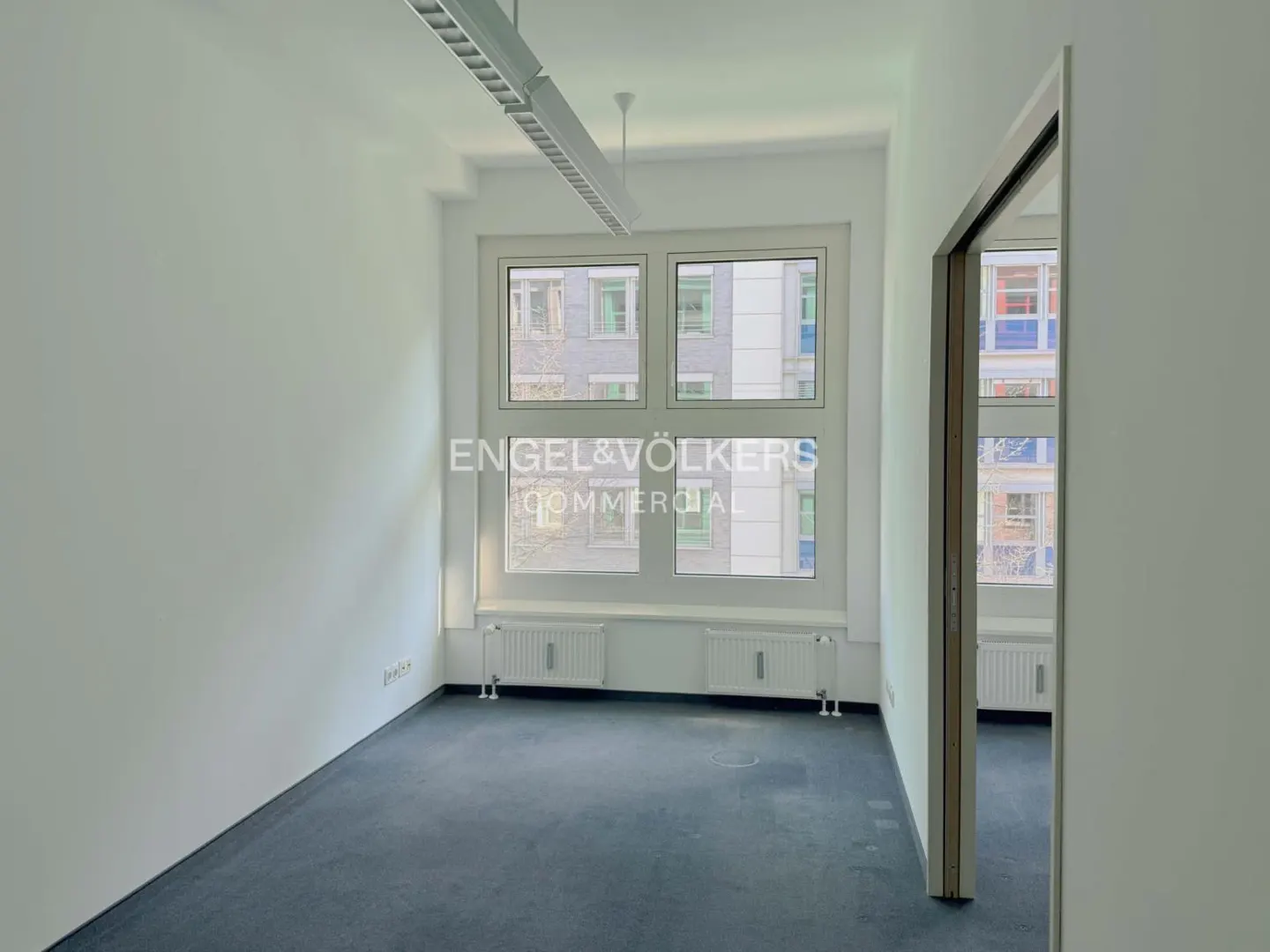 Empty office space with blue carpet, white walls, and a large window. A doorway is visible on the right.