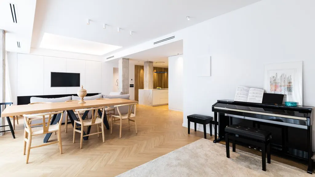 Bright, modern living space with a dining table, piano, and open kitchen. White walls and light wood floors create a spacious feel.