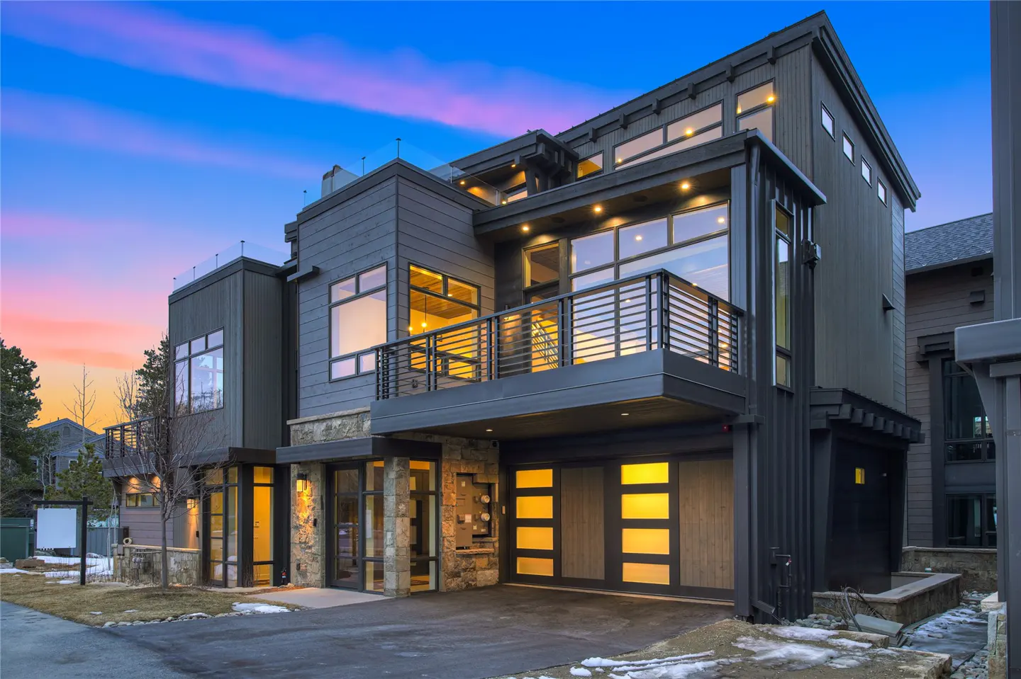 Modern gray house with a balcony and large windows, lit from within, at dusk.