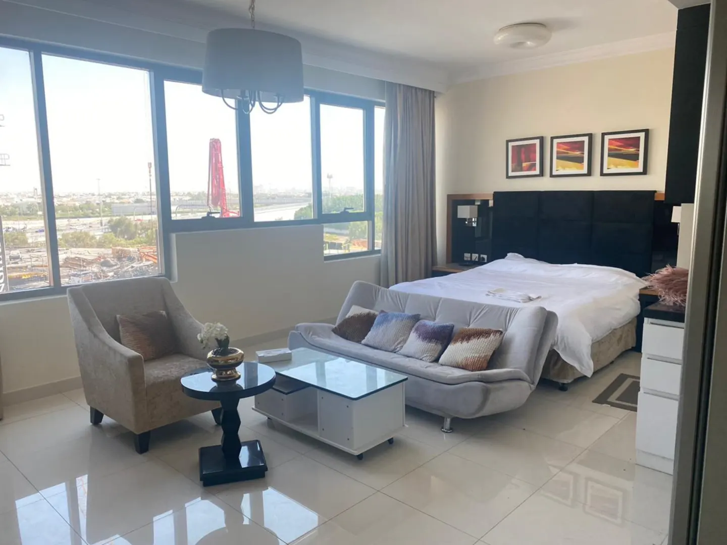 Bright studio apartment with a beige armchair, grey sofa, and a bed with a black headboard. Large windows offer a city view.