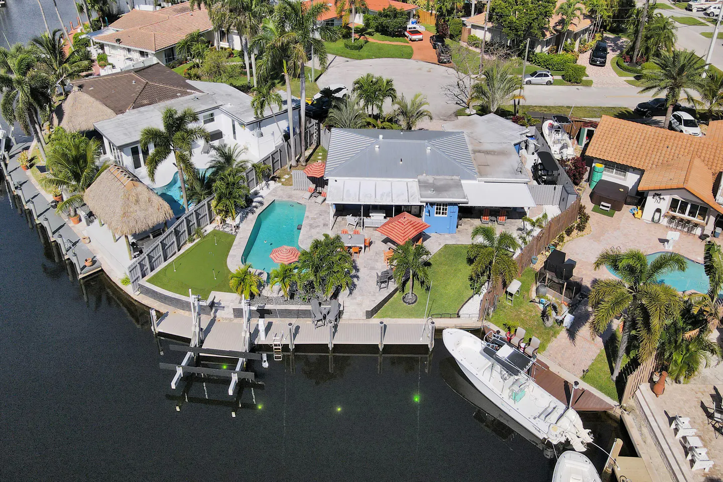 Aerial view of waterfront homes with pools, docks, and boats on a sunny day. Palm trees and green lawns surround the properties.