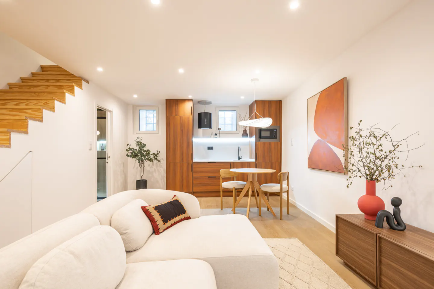 Bright studio apartment with white walls, wood floors, and a small kitchen. A white sofa sits in the foreground, with a wooden staircase to the left.