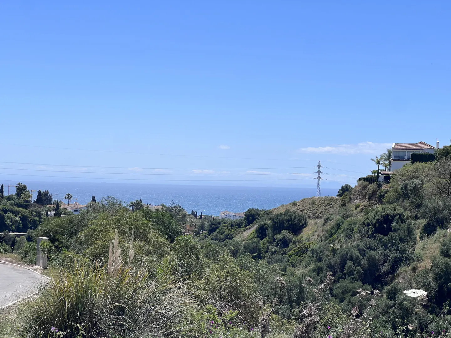 Scenic view of the ocean and a hillside with lush green vegetation under a clear blue sky. A house sits atop the hill.
