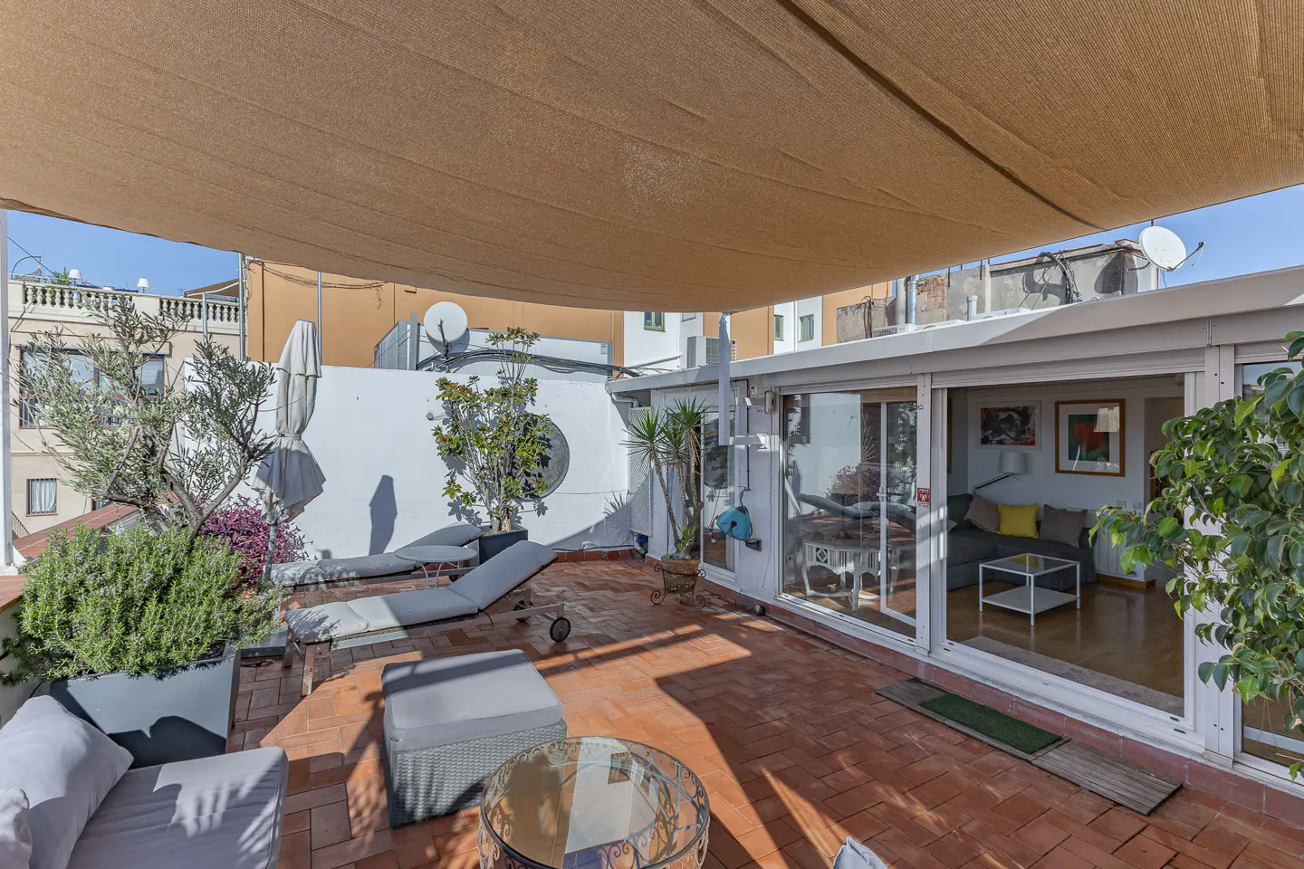 Rooftop patio with brick flooring, lounge chairs, and a glass table. A beige sunshade covers the area. Sliding glass doors lead to an indoor living space.