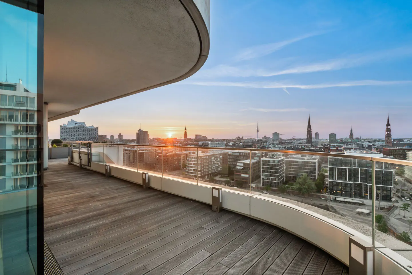 A curved wooden deck with a glass railing overlooks a city skyline at sunset. The sky is blue and orange.
