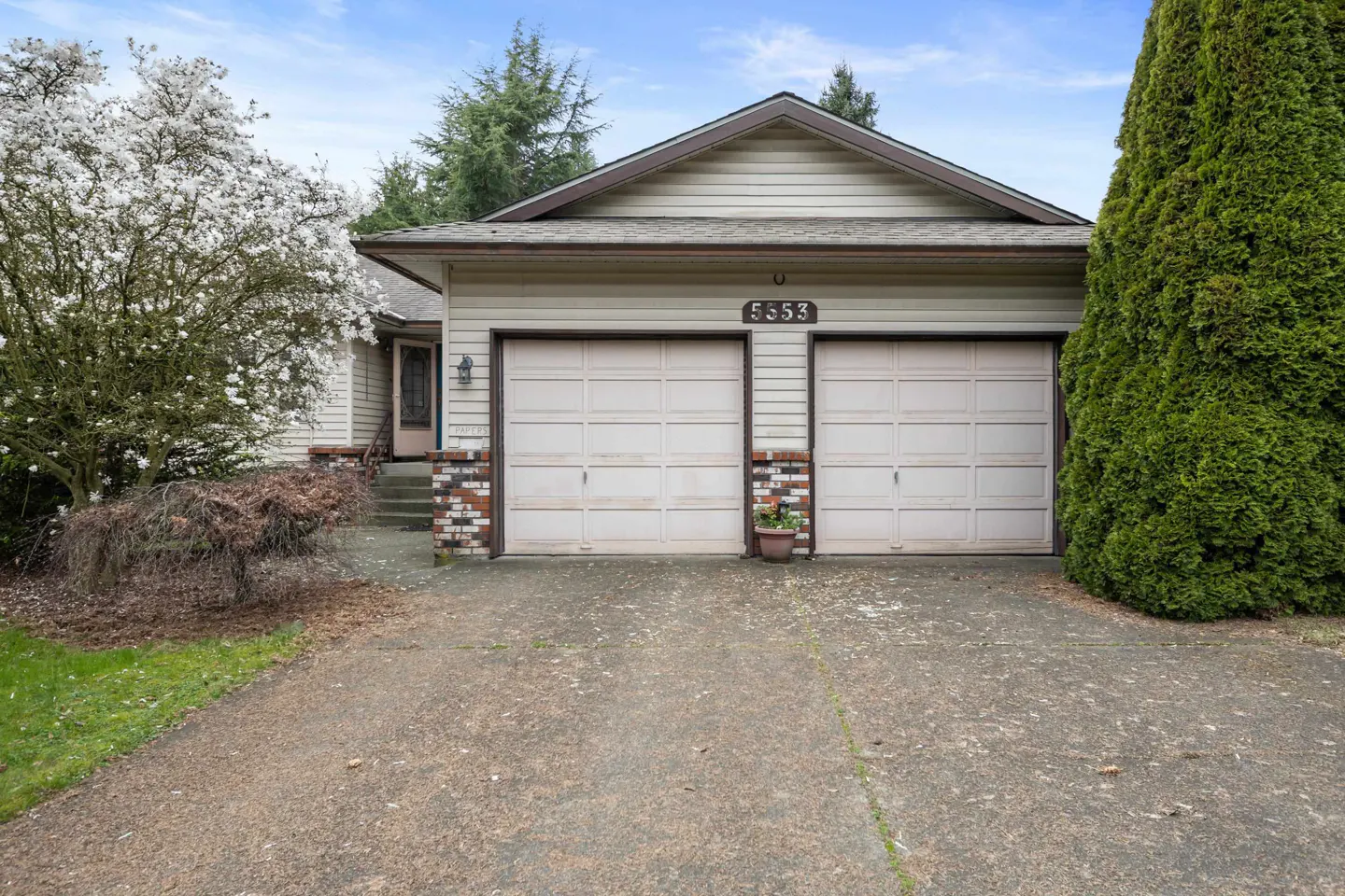 Tan house with a two-car garage, concrete driveway, blooming white tree, and tall evergreen on a sunny day.