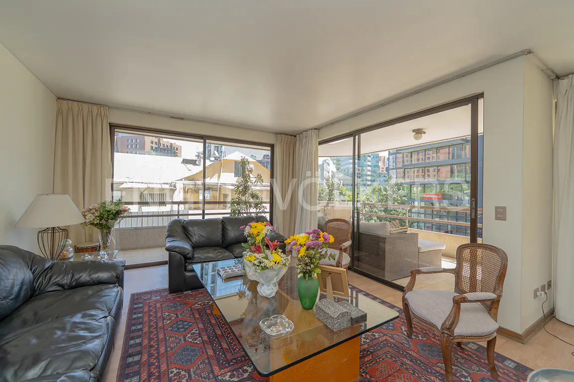 Living room with black leather sofas, glass coffee table with flowers, and a red patterned rug. Balcony with city views visible through sliding glass doors.