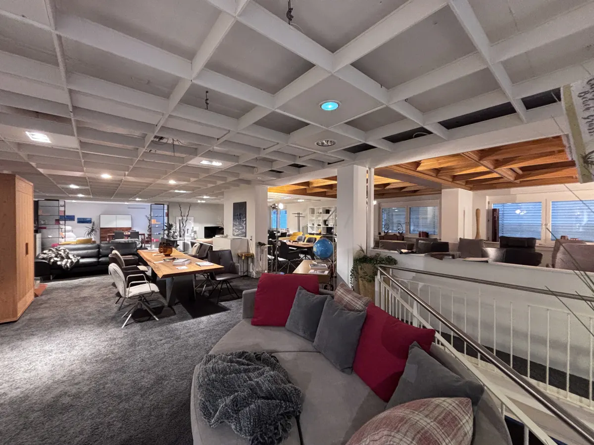 A furniture showroom with a gray carpet, a gray sofa with red pillows, and a wooden table with chairs.