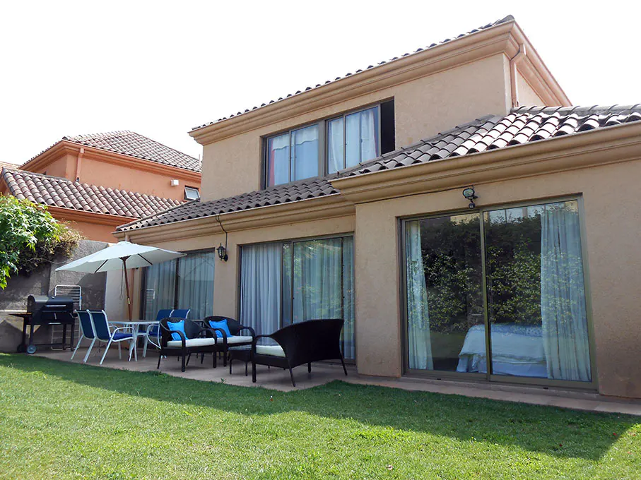 Backyard view of a two-story beige house with a patio set, grill, and green lawn.