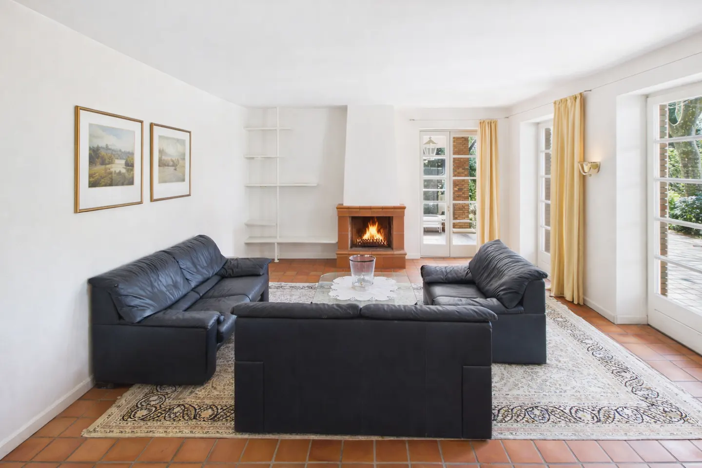 Bright living room with black leather sofas, a fireplace with a burning fire, and French doors leading to a patio.