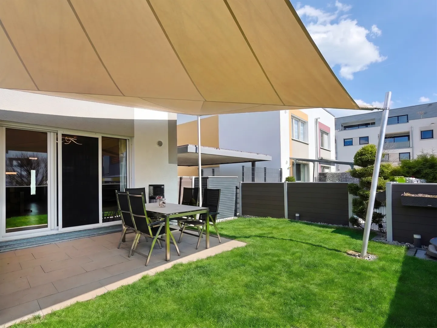 Outdoor patio with a beige sunshade, a table with six chairs, and a green lawn. Modern houses are in the background.