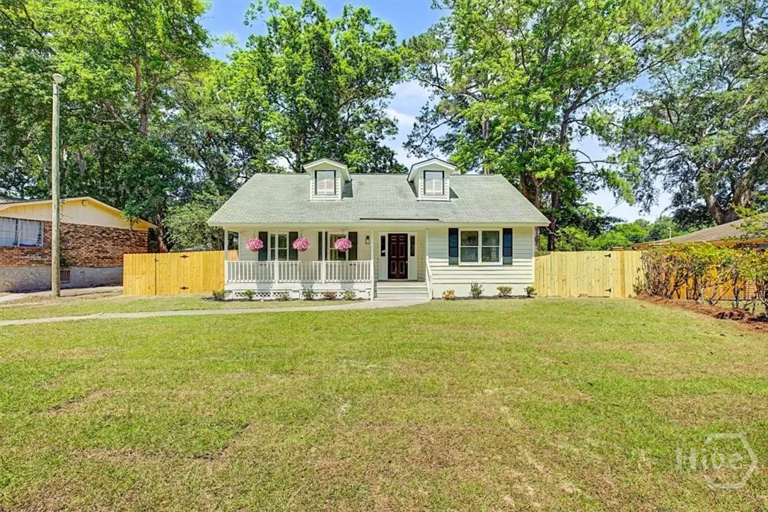 A white, one-story house with a green roof, black shutters, and a white porch with pink flowers. A green lawn and wooden fence surround the house.