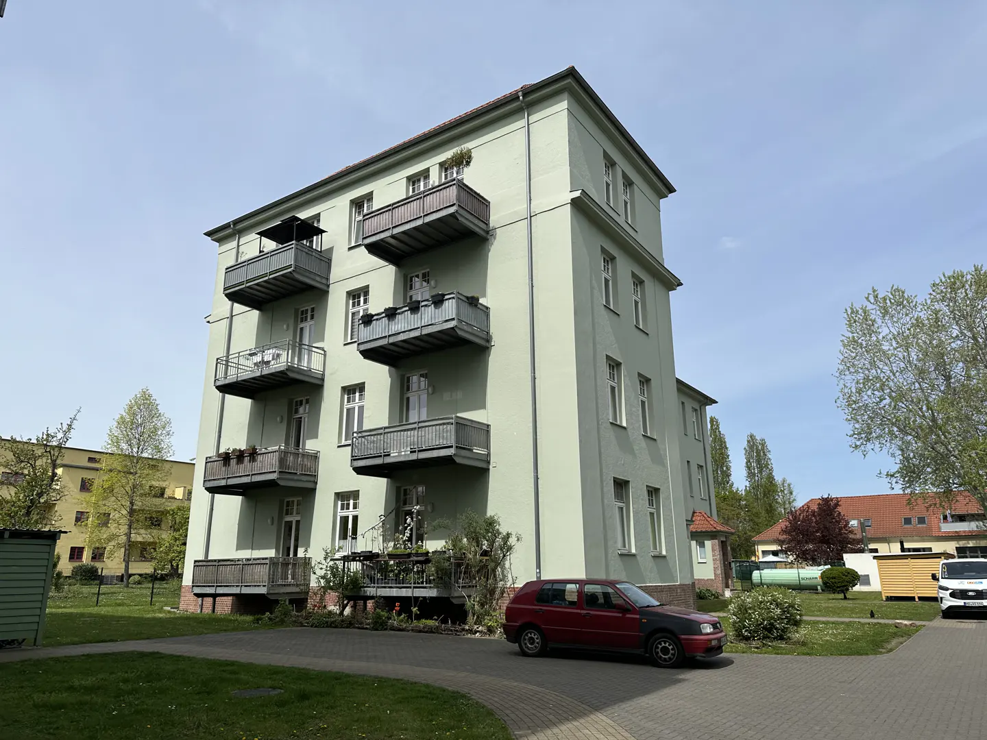 A light green apartment building with balconies and a red car parked in front.