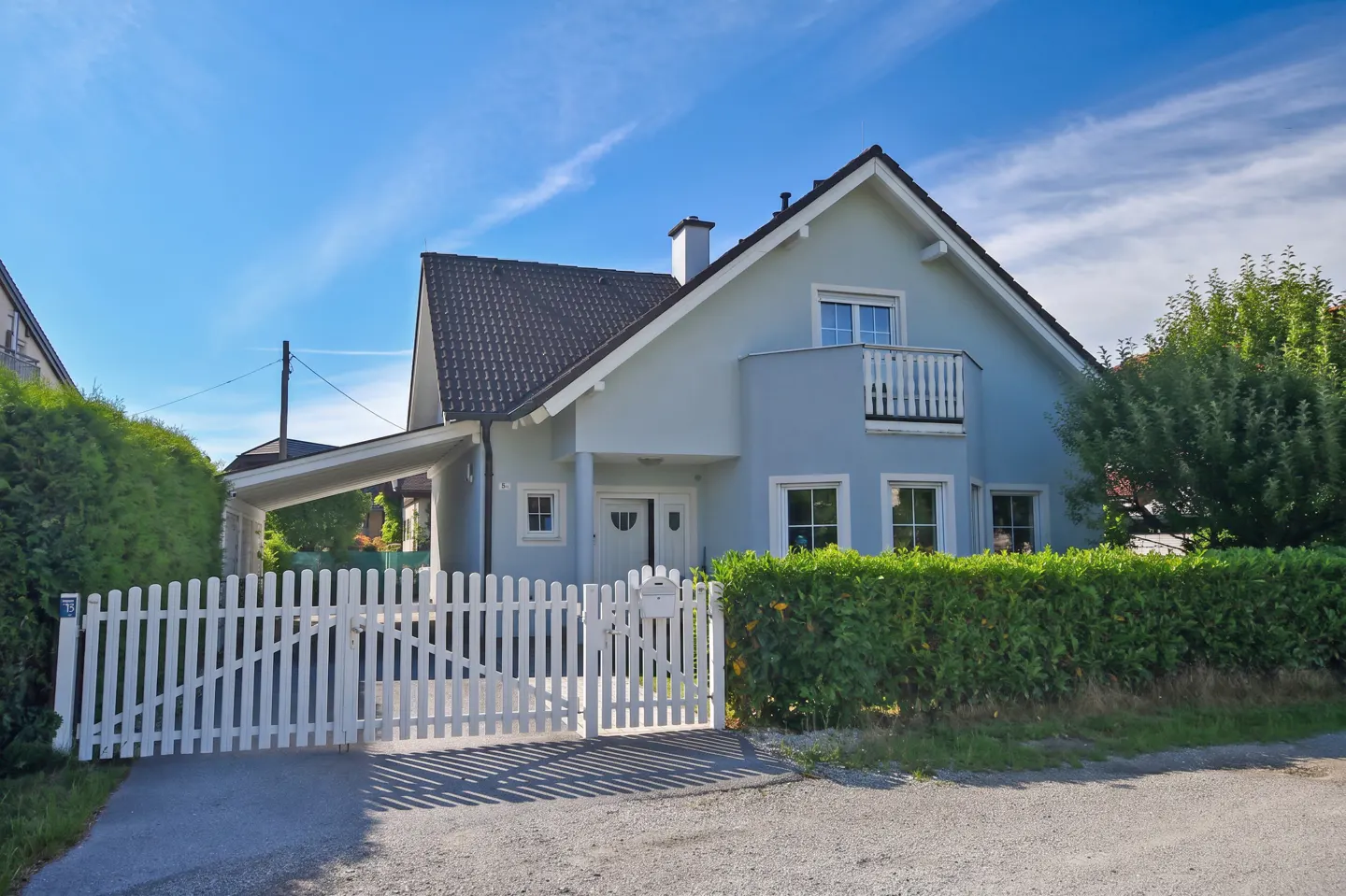 Light blue two-story house with a dark roof, white picket fence, and attached carport under a bright blue sky.