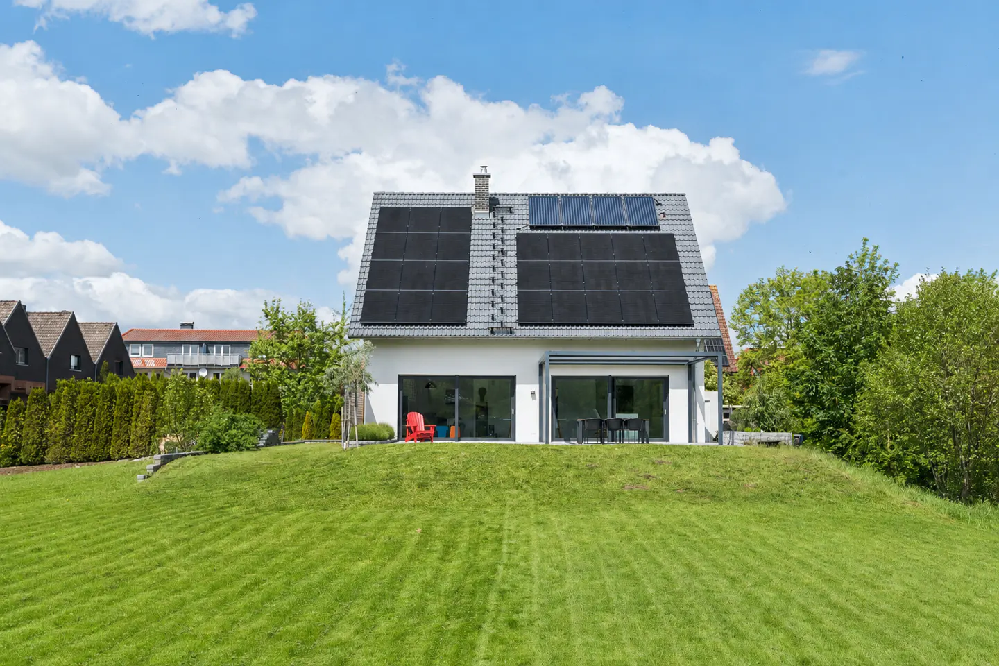 Modern white house with solar panels on a grey roof, large glass doors, and a red chair on a lush green lawn.