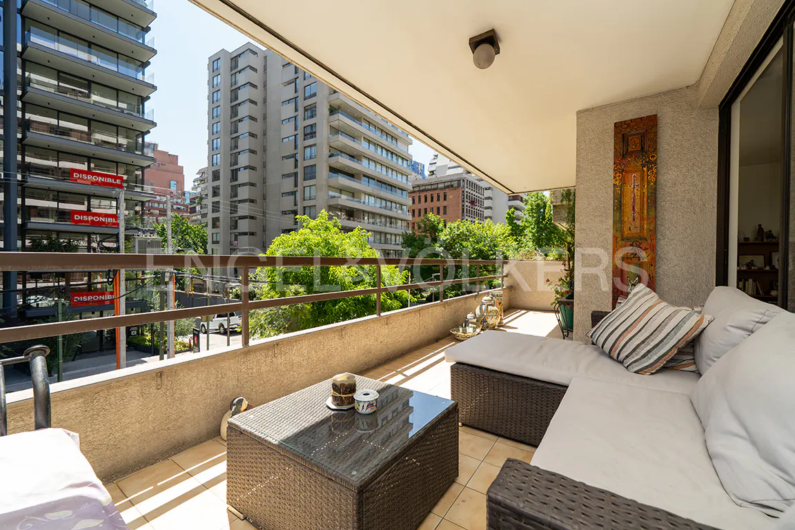 Balcony view with wicker furniture, including a table and sectional sofa with white cushions. City buildings and green trees are visible in the background.