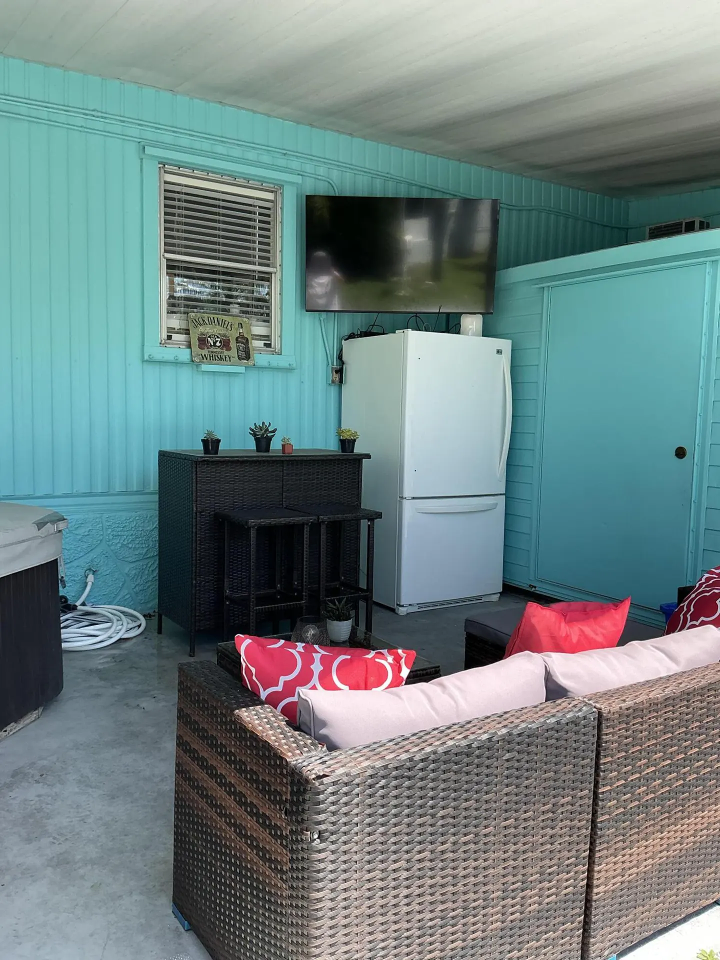 Covered patio with turquoise walls, wicker sofa with red pillows, bar stools, and a white refrigerator with a mounted TV.