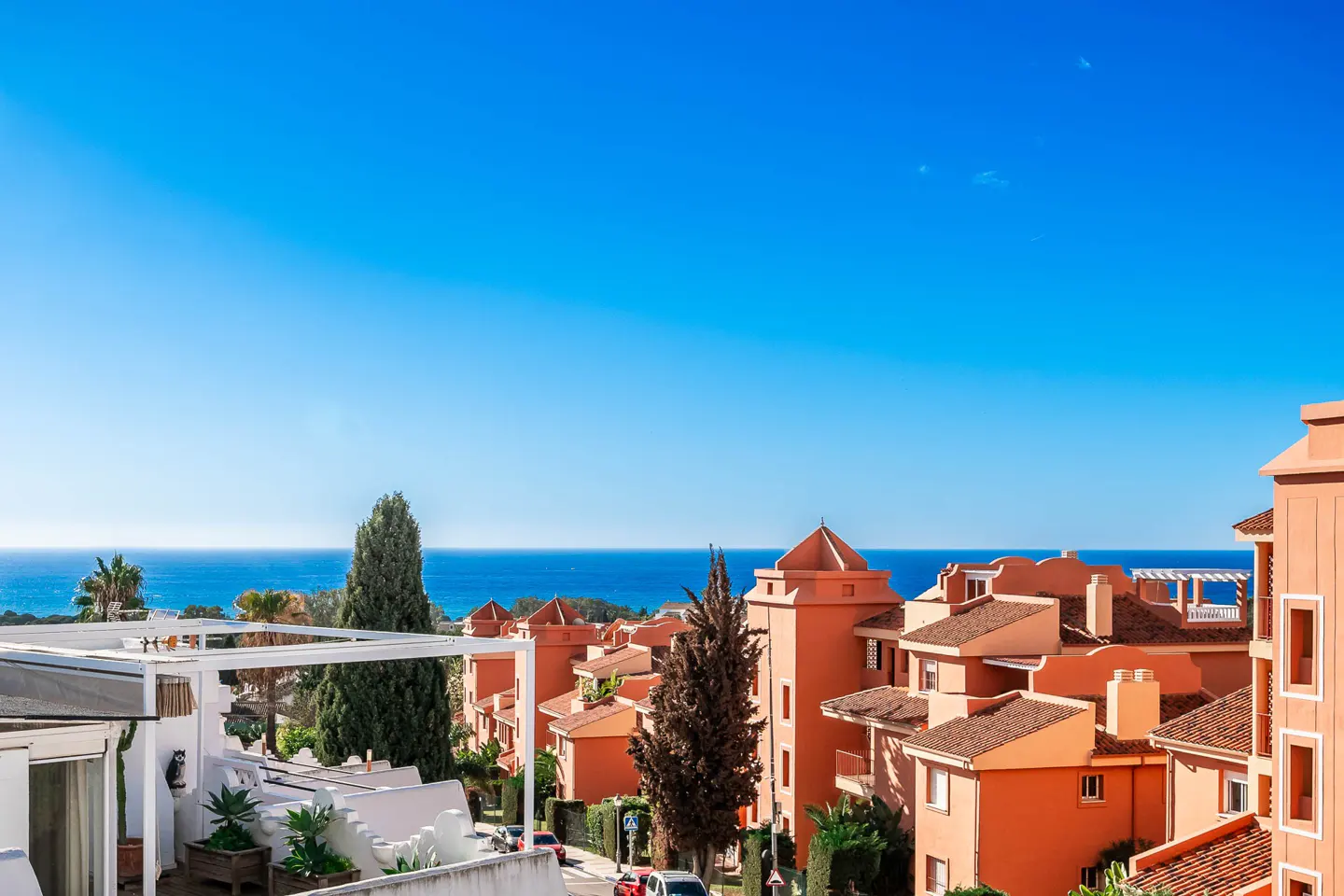 View of orange-colored buildings with red tile roofs, trees, and the ocean under a clear blue sky.