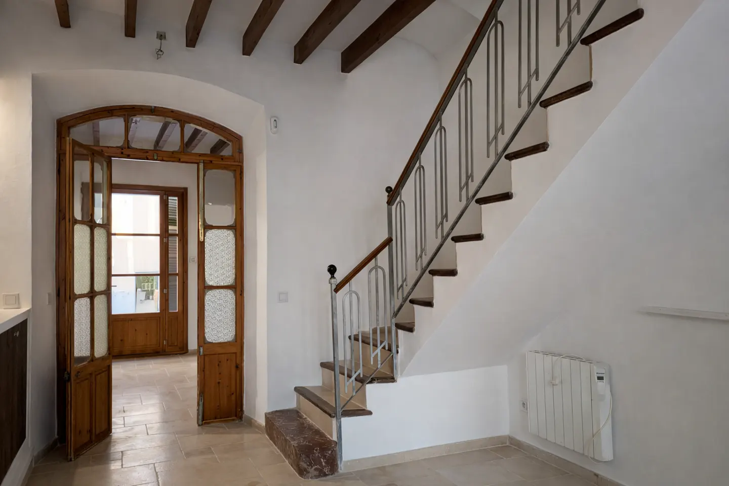 Interior view of a white-walled foyer with a wooden staircase and an arched wooden door. The floor is tiled, and wooden beams are visible on the ceiling.