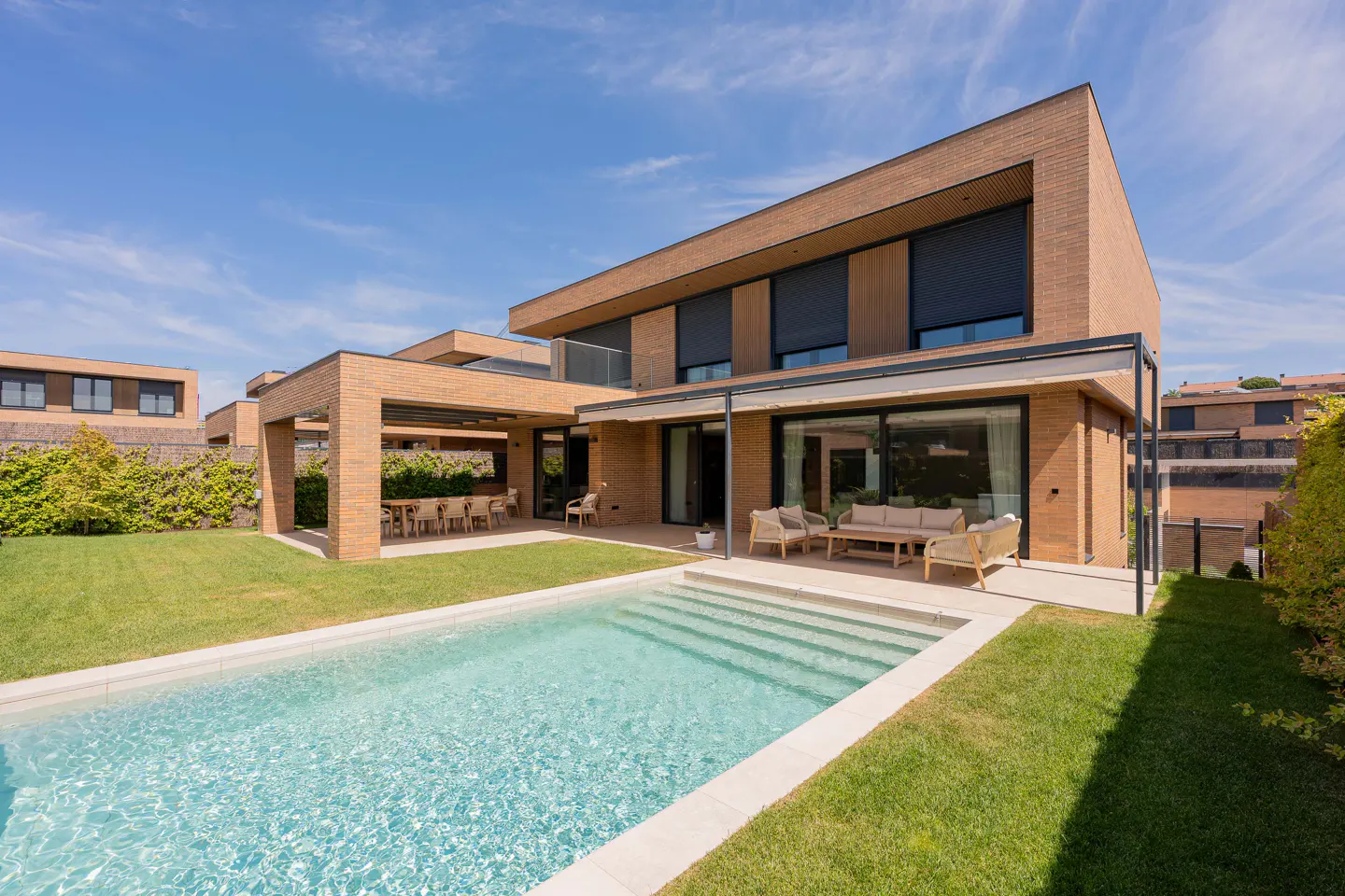 Modern brick house with a pool, patio furniture, and dining area on a sunny day.