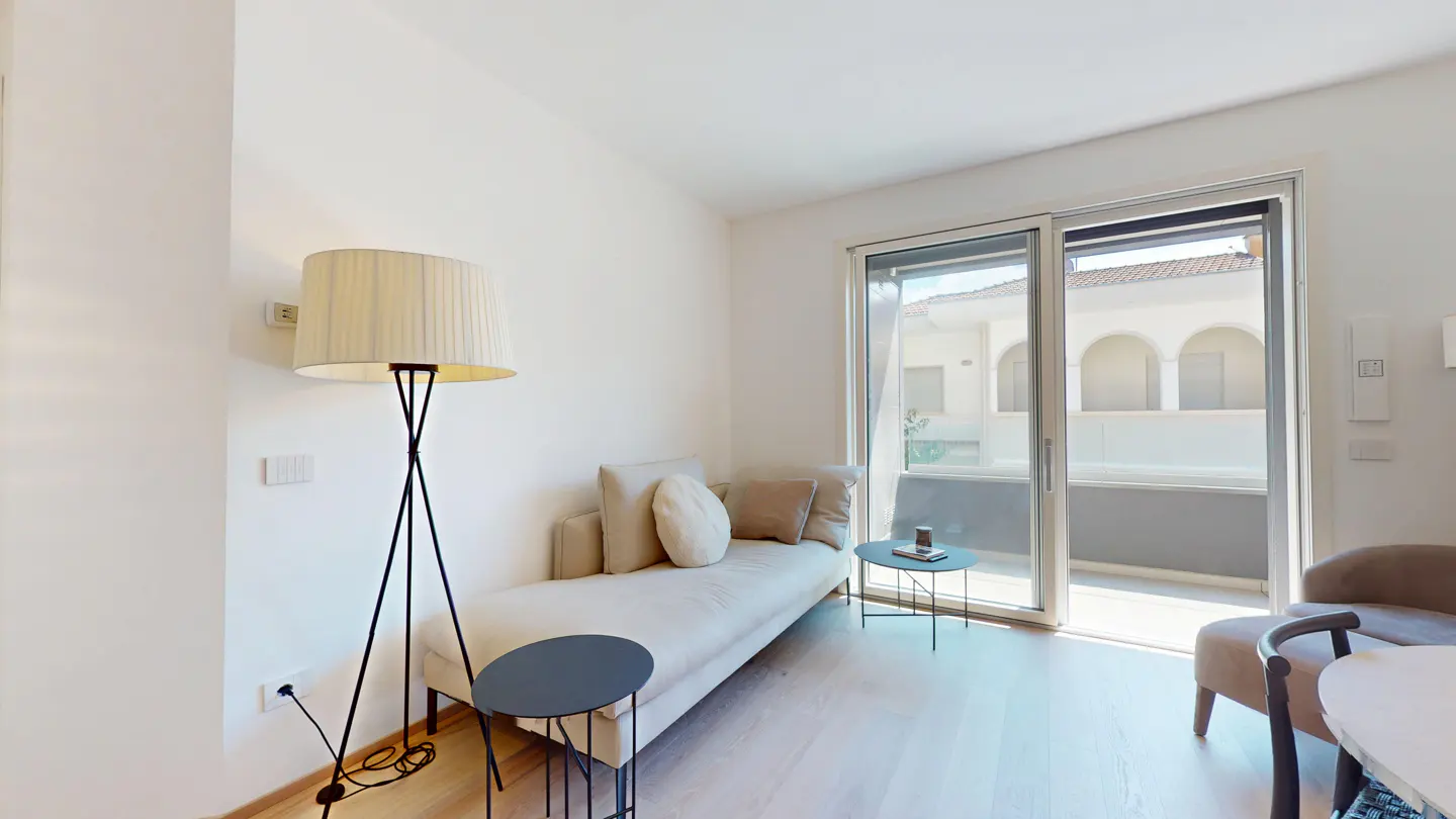 Bright living area with a beige daybed, floor lamp, small side tables, and a sliding glass door leading to a balcony.