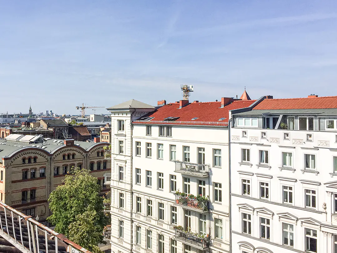 Cityscape view of Berlin, Germany, featuring white buildings with red tile roofs under a blue sky. Balconies and a construction crane are visible.