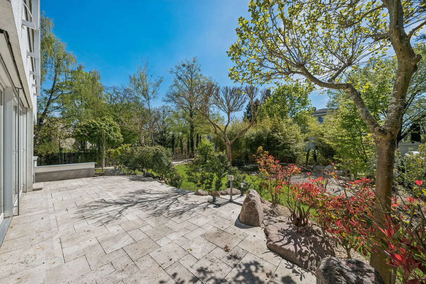 Stone patio with lush garden. Trees, red shrubs, and rocks surround the patio under a bright blue sky.