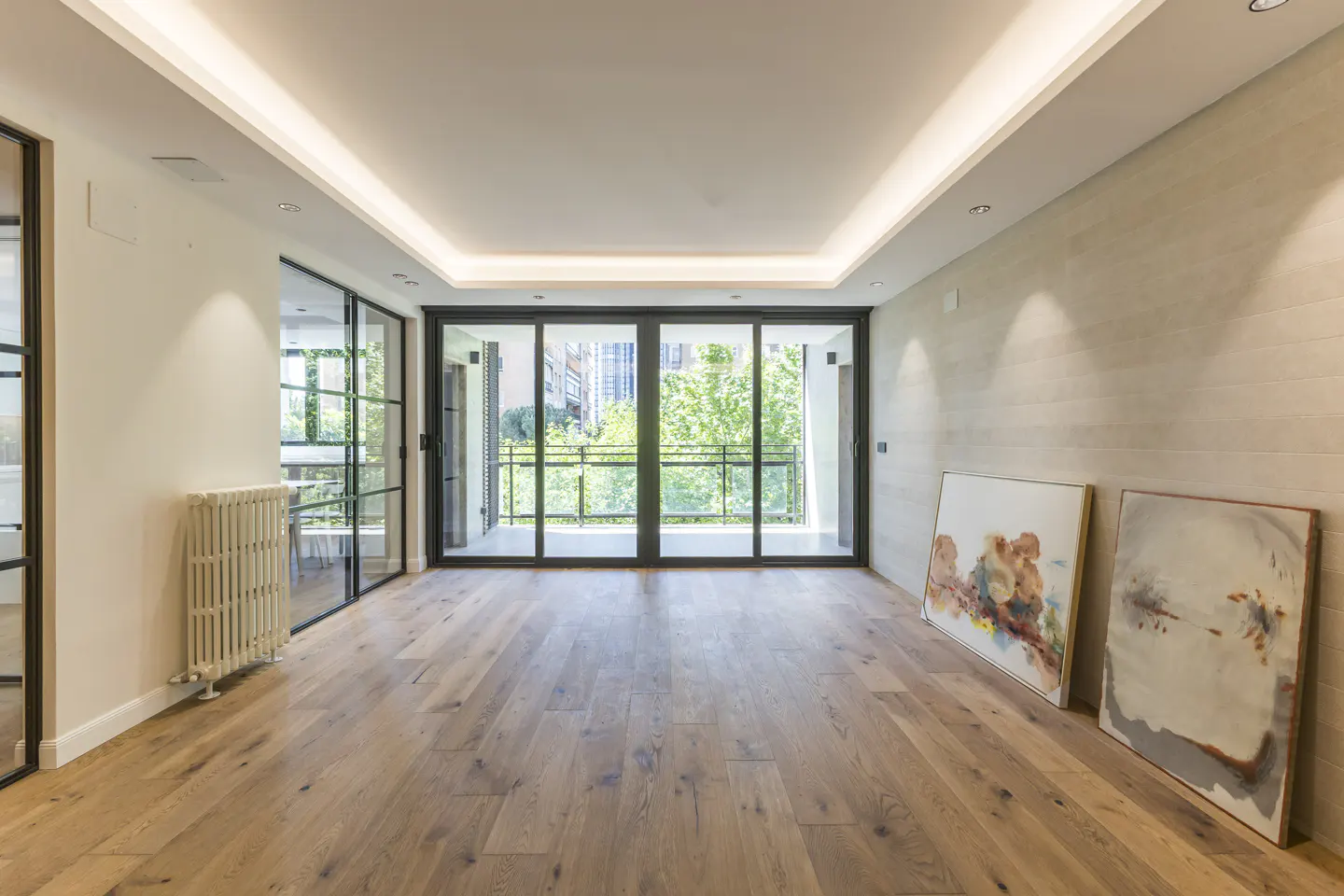 Bright, empty room with wood floors, white walls, and black-framed glass doors to a balcony with green trees visible outside. Two paintings lean against the wall.
