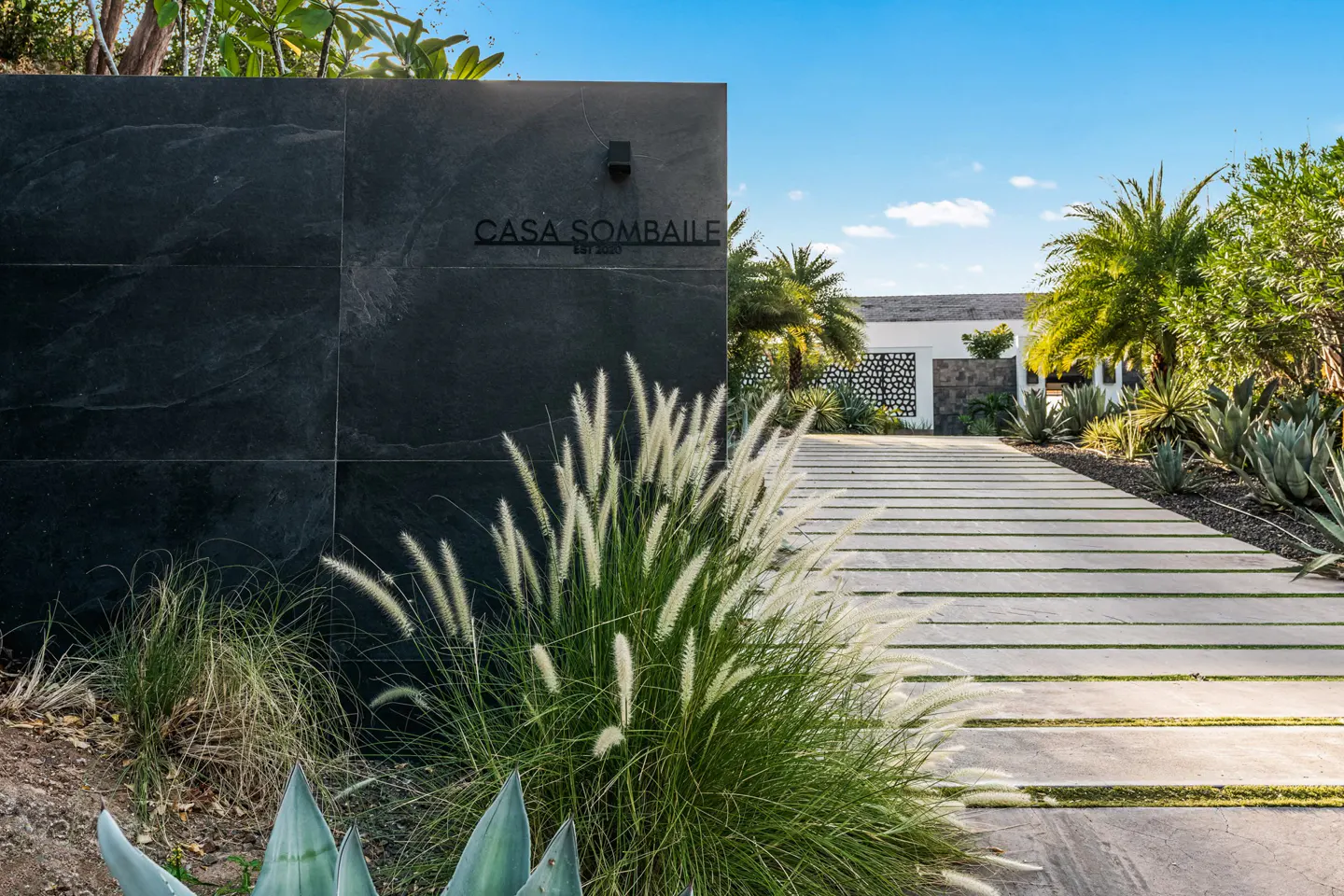 Exterior shot of Casa Sombaille, featuring a stone wall with the house name, a concrete driveway with grass, and tropical plants.
