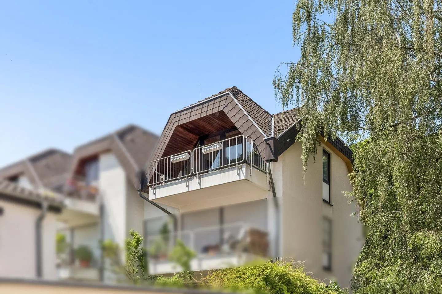 Exterior view of a white residential building with a brown tiled roof, featuring a balcony and a large birch tree.