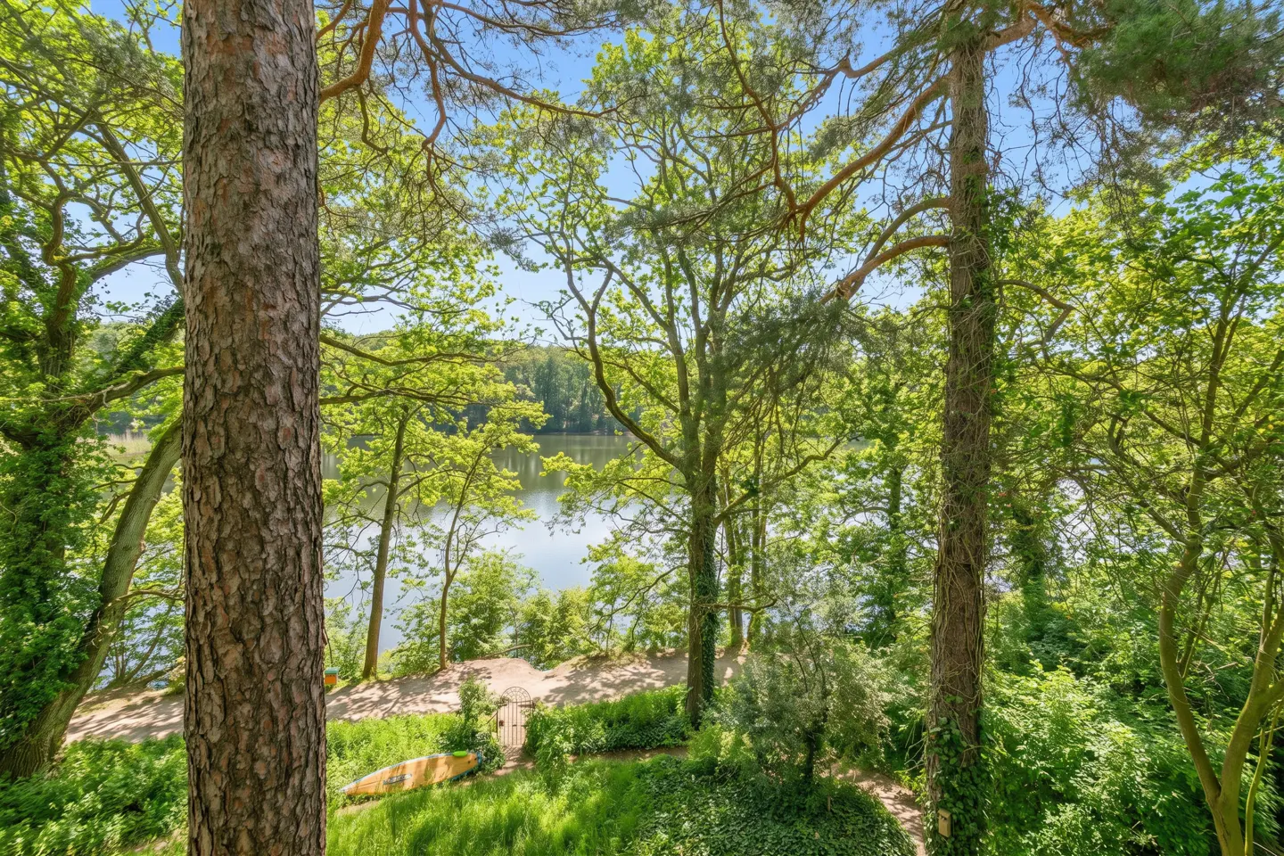 Lush green trees frame a lake view. A yellow kayak rests on the grass near a gate leading to the water.