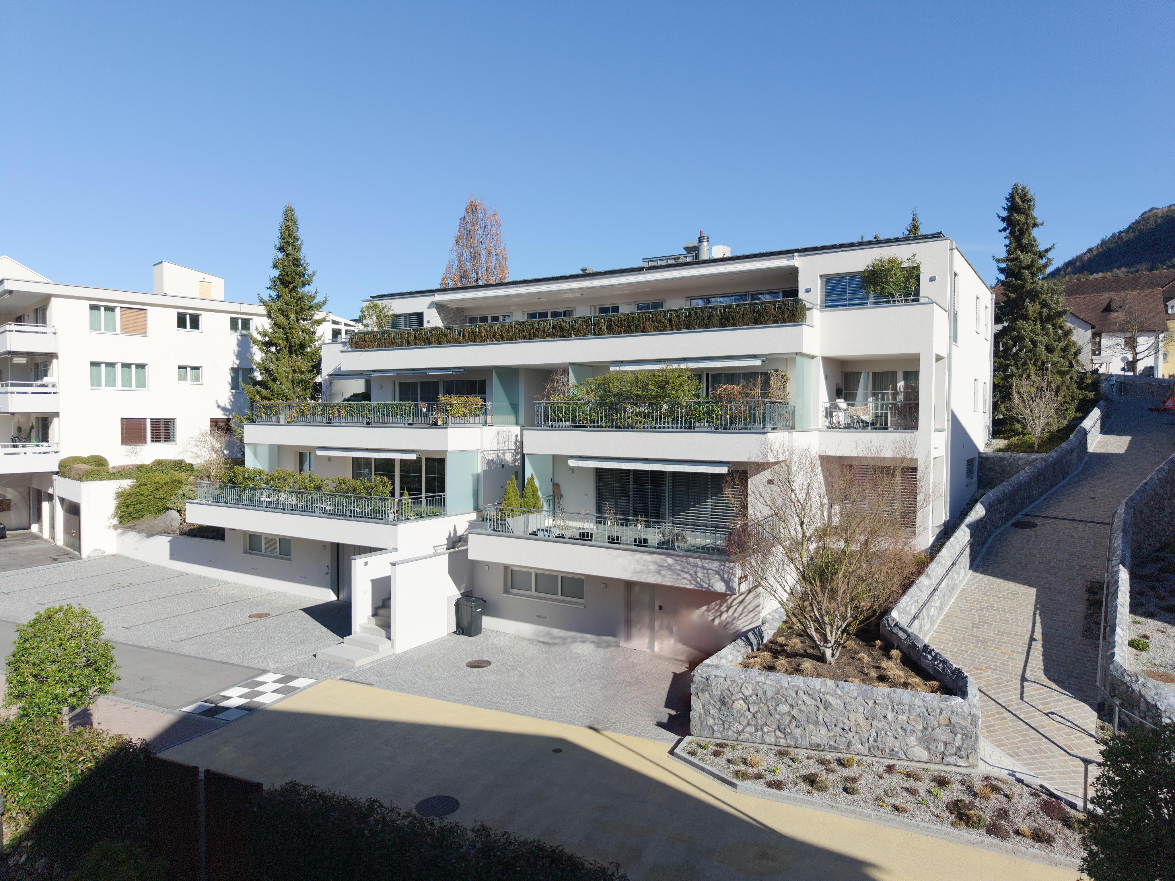 Modern white multi-story apartment building with tiered balconies, landscaping, and a paved driveway under a clear blue sky.