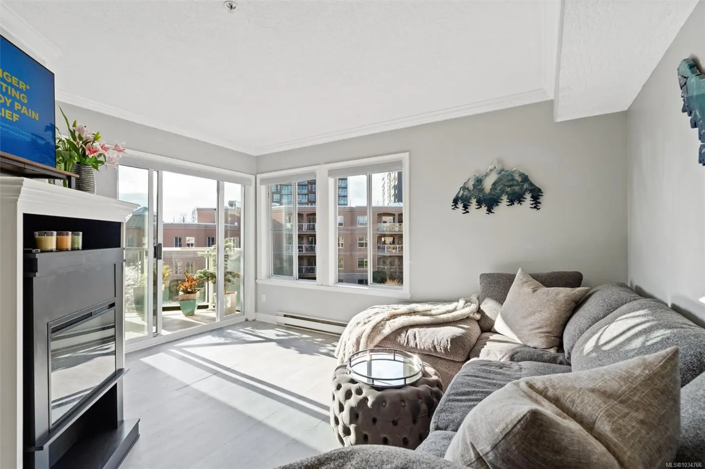 Bright living room with gray sectional sofa, ottoman, and mountain wall art. Sliding glass doors lead to a balcony with plants.