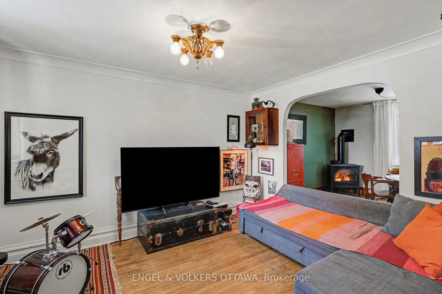 Living room with a drum set, donkey art, TV on a trunk, and a wood-burning stove in the next room.