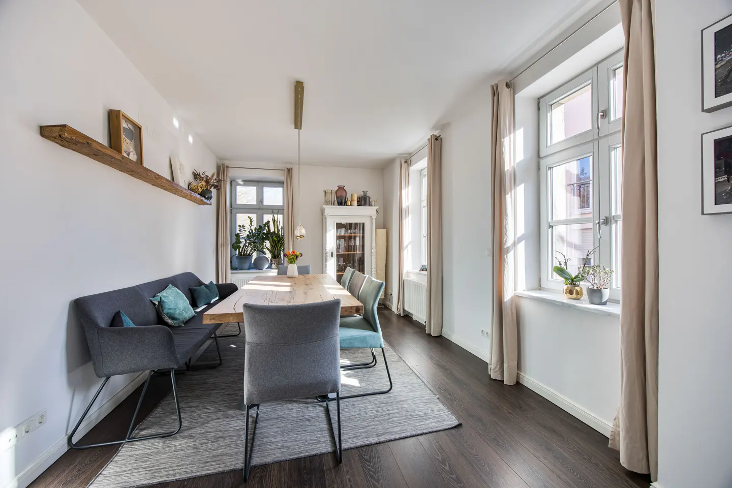 Bright dining room with a long wooden table, gray chairs, and a bench with teal pillows. Windows with beige curtains let in sunlight.