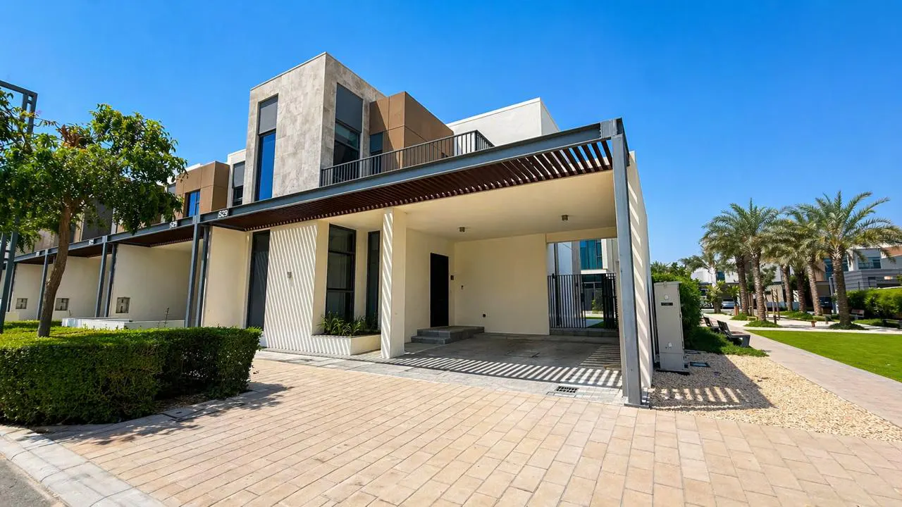 Modern townhouse with a carport, featuring a beige brick driveway, white walls, and a brown accent wall under a clear blue sky.