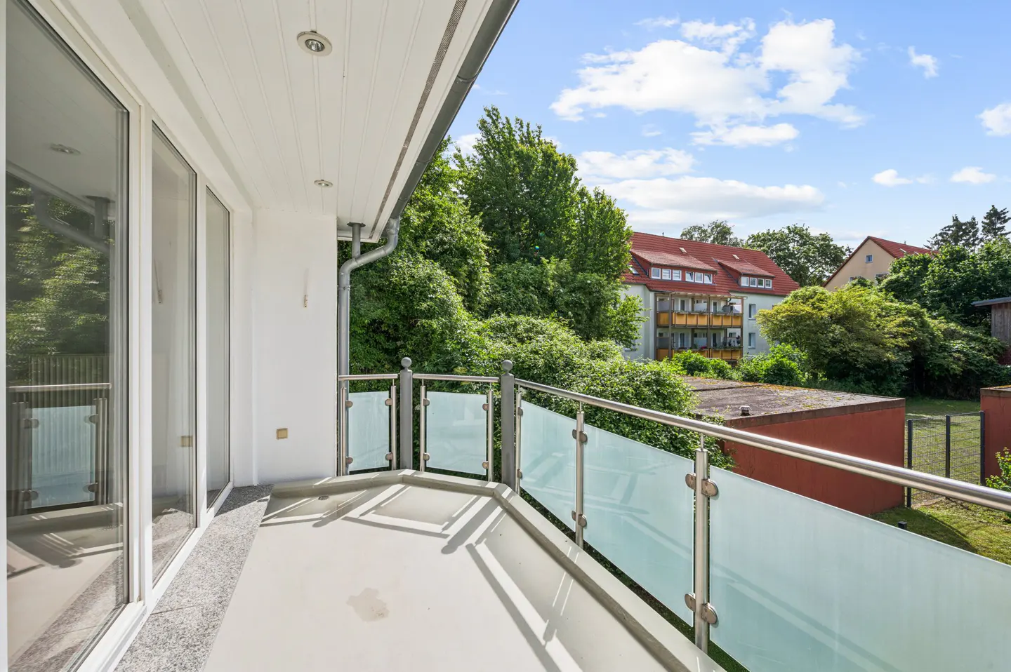 Modern balcony with glass railing, white ceiling, and view of lush green trees and neighboring residential buildings.