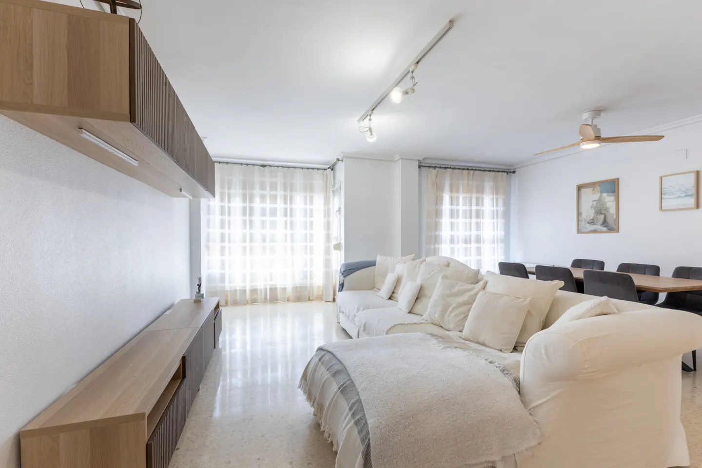 Bright living room with white sofa, pillows, and blanket. A wooden entertainment center and dining table are visible.