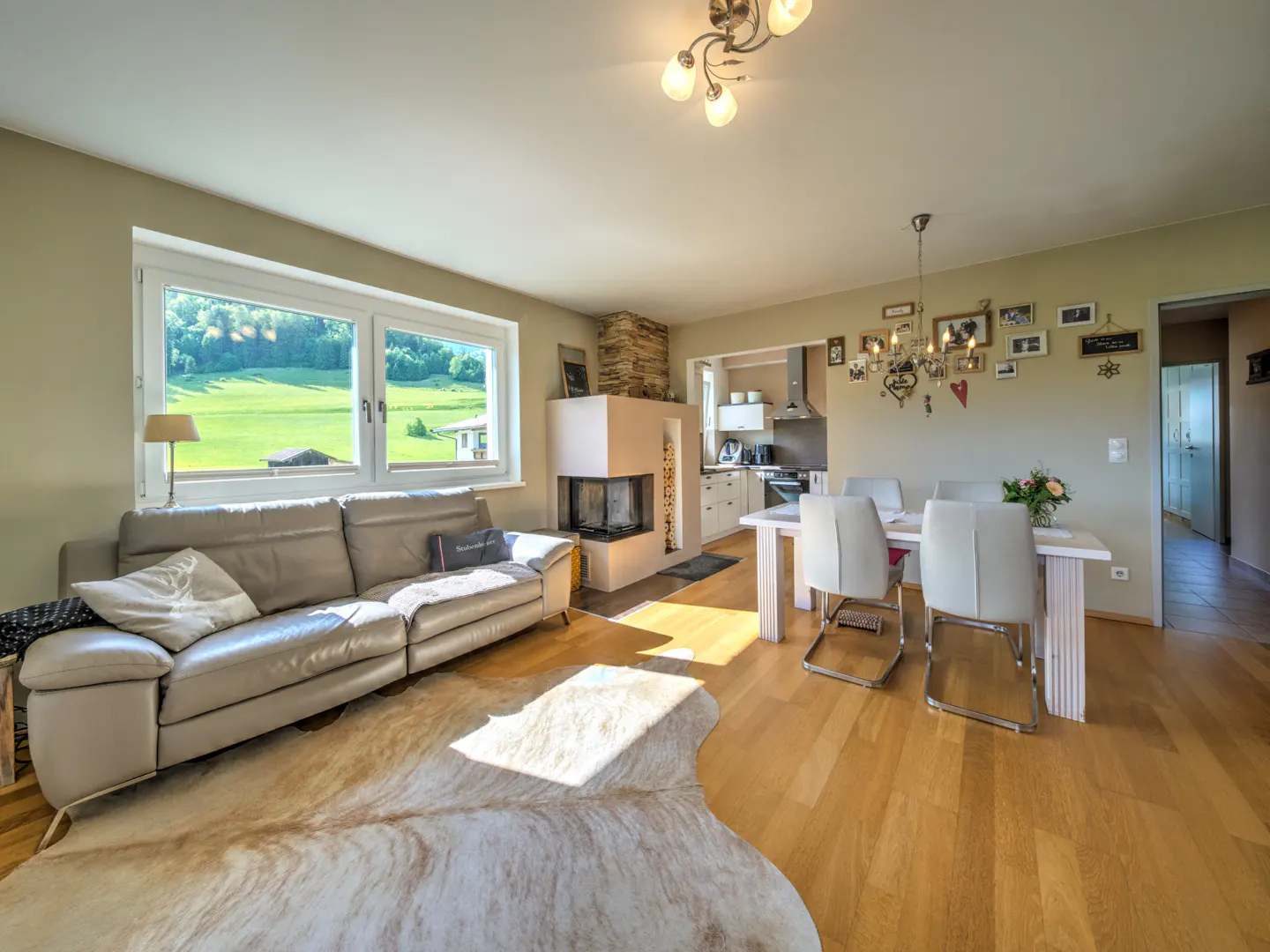 Bright living room with a beige sofa, fireplace, dining table, and a view of green fields through a large window.