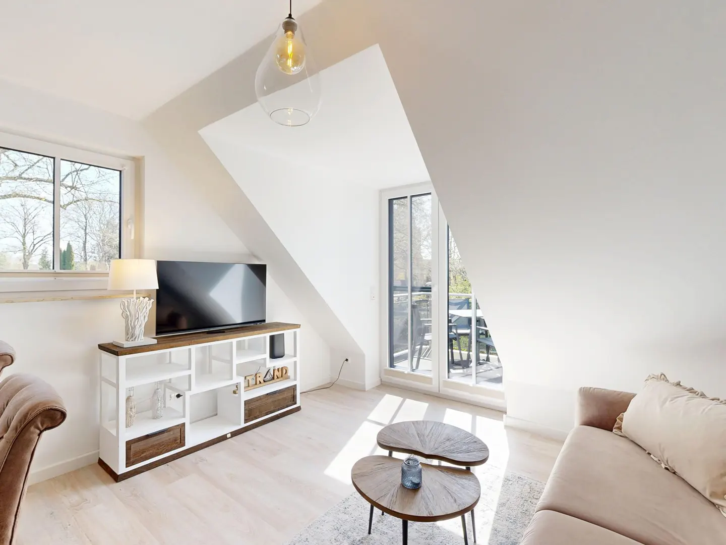 Bright attic living room with a beige sofa, two wooden coffee tables, a TV on a white media console, and a balcony door.