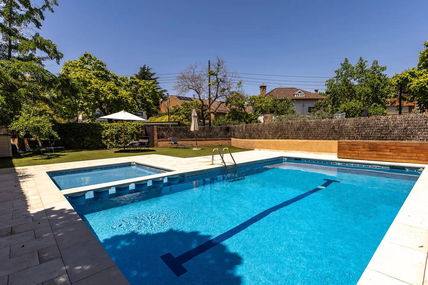 A bright blue swimming pool with a lap lane, stone patio, and green lawn with lounge chairs under a white umbrella.