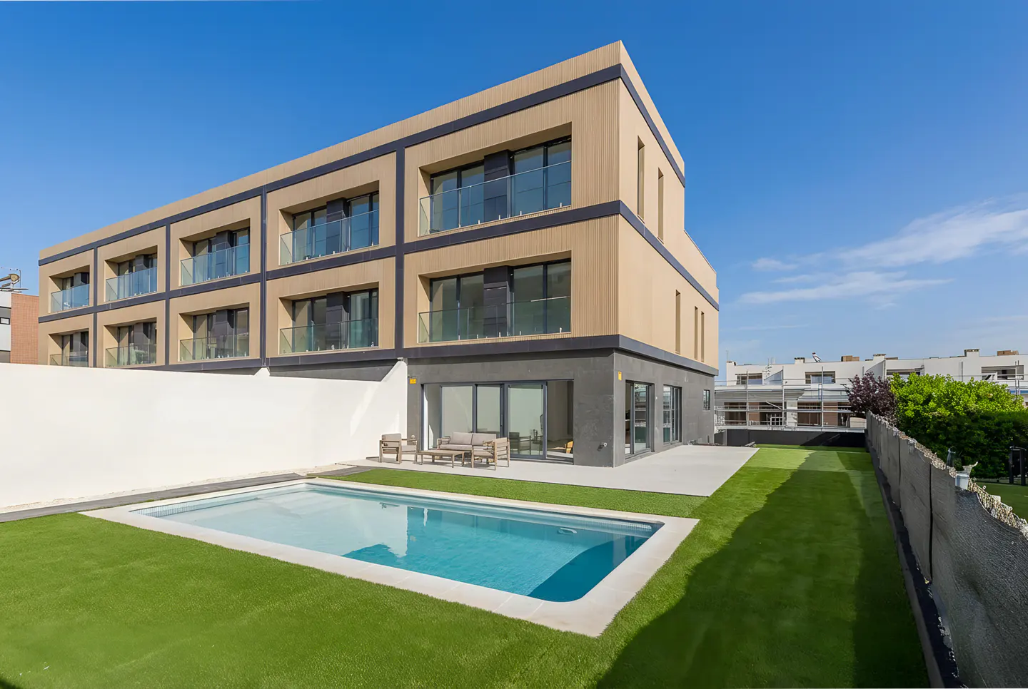 Modern three-story building with a pool and artificial grass lawn under a blue sky. Balconies with glass railings are visible.