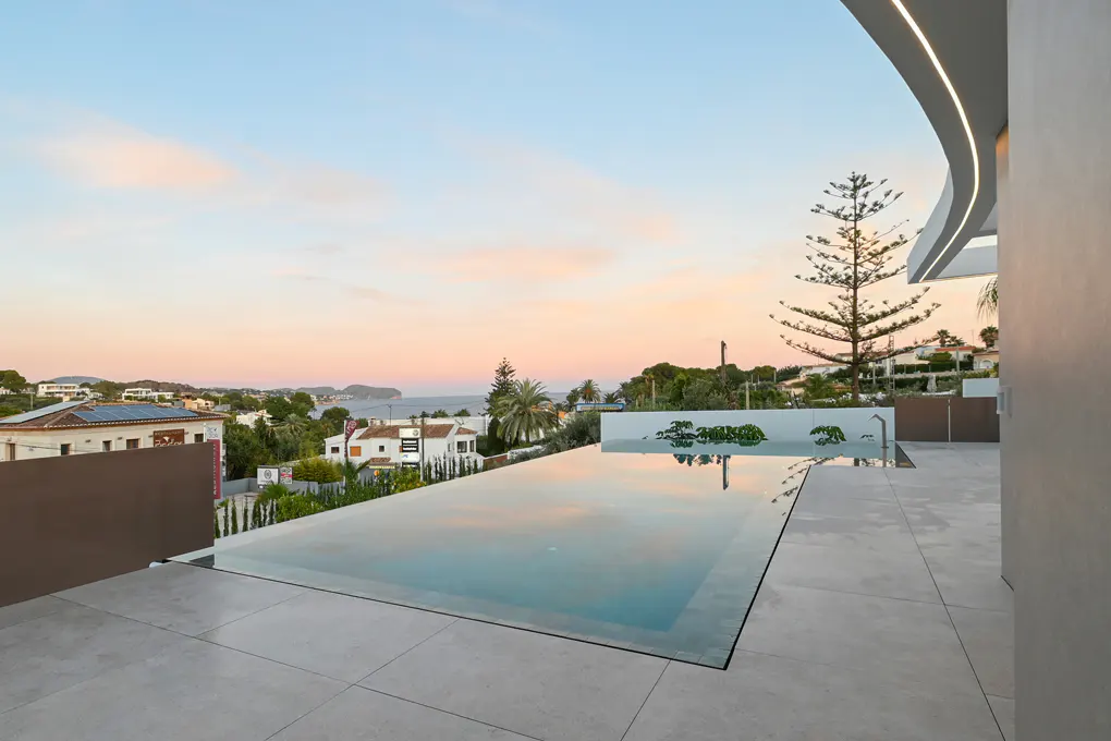 Infinity pool on a tiled patio overlooks a coastal town at sunset. The sky is pink and blue.