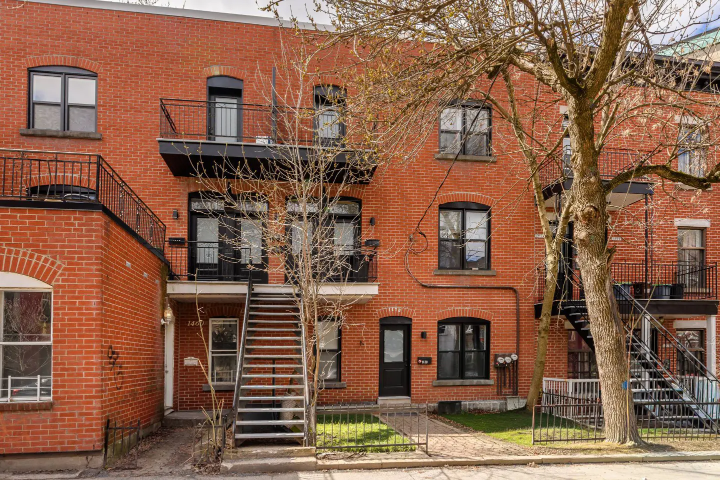 Red brick apartment building with black metal stairs and balconies. Trees in front of the building.