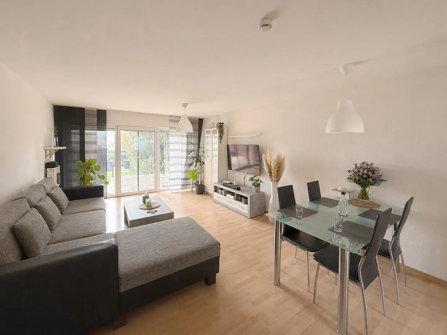 Bright, modern living room with a gray sectional sofa, glass dining table with black chairs, and sliding glass doors to a garden.