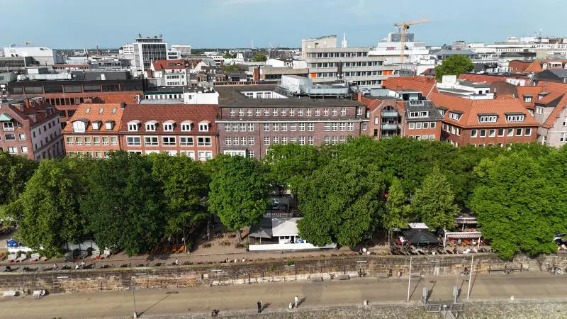 Aerial view of a city waterfront with a stone wall, lush green trees, outdoor cafes, and historic brick buildings behind.
