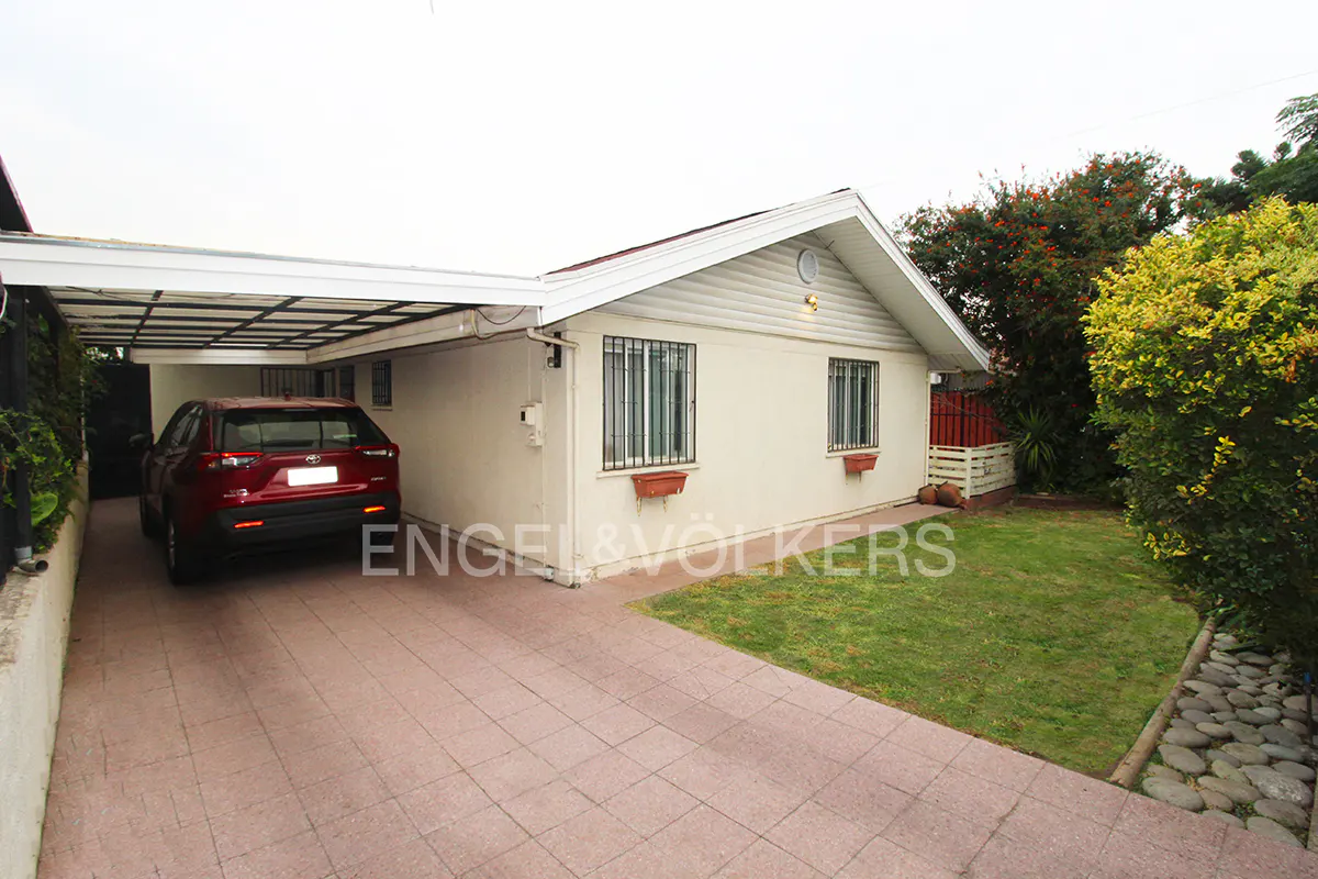 Exterior view of a one-story house with a red car parked in the driveway. The house is light beige with a white roof and trim. There is a small lawn to the side of the house.