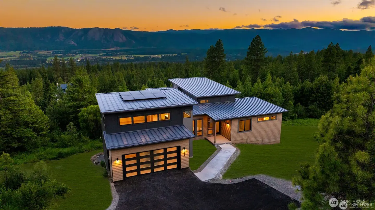 Modern home with a metal roof and solar panels, nestled in a forest with mountains in the background at sunset.