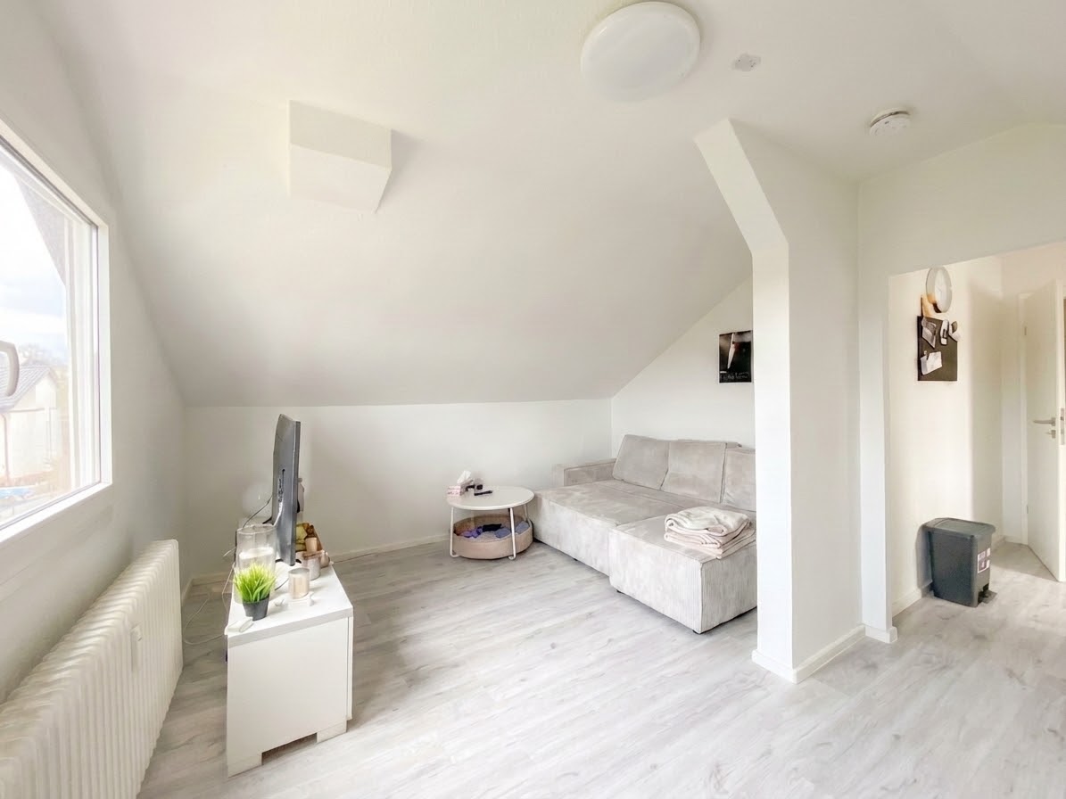 Bright, modern attic living room with white walls, light wood floors, and a gray sectional sofa. A TV sits on a white console table.