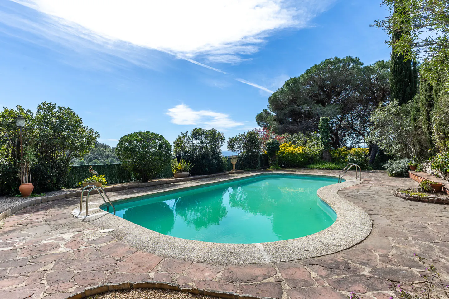 Outdoor pool with turquoise water, surrounded by stone patio and lush greenery under a blue sky.